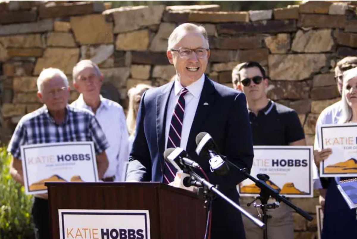 John Giles speaking a podium with two microphones in front of him. Behind him is a group of Katie Hobbs supporters holding her campaign signs.